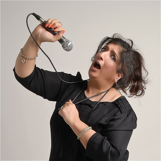 South Asian female comedian wearing black top with long dark hair in ponytail with mic lead wrapped around neck as if being strangled whist holding mic in right hand about to speak - face looks faux worried.