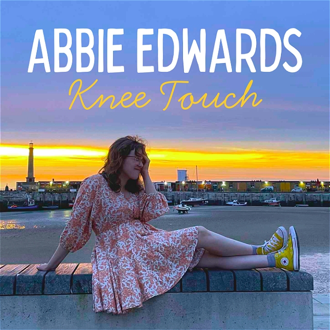 Female comedian with brown hair and glasses sitting on a beach wall in Margate in front of a sunset with her hand on her forehead