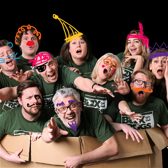 Many Improvisers with colourful hats and glasses smile and laugh from inside a cardboard box
