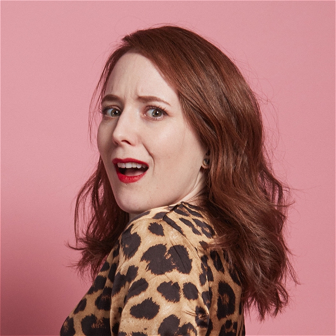 A comedian wearing a leopard print dress pulls a face in front of a pink background.