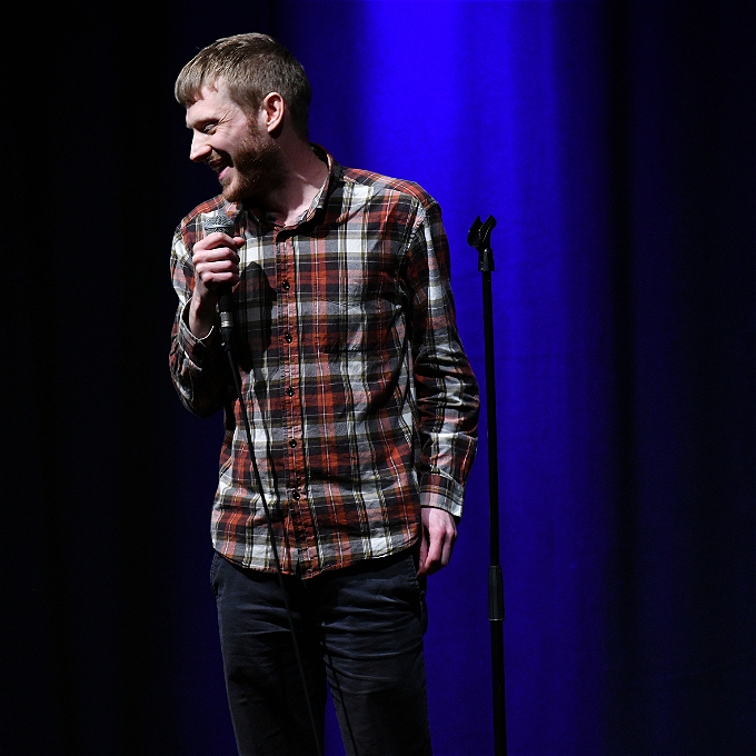 David Eagle in a checked shirt smiling and holding a microphone in front of blue curtains