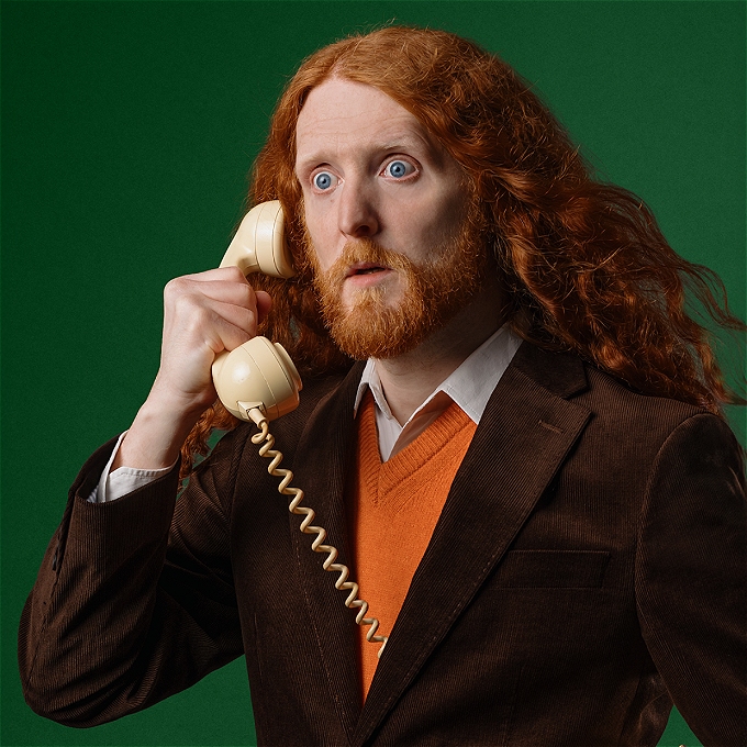 A man with long ginger hair and a beard looks shocked answering an old wired telephone emerging from inside his suit