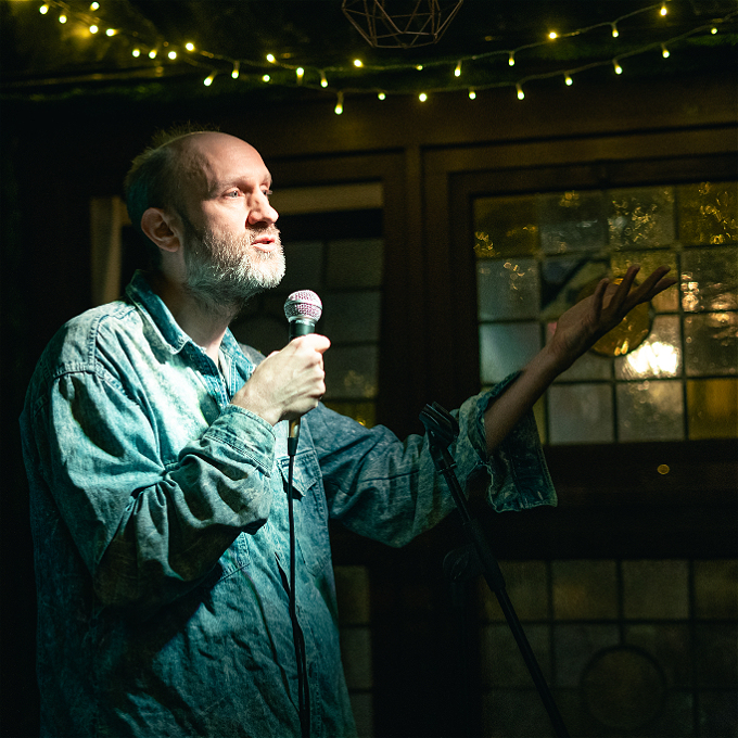 A heavily lit man in a speckled sky blue denim jacket, bald, mid 40s, with a greying beard, speaks into a microphone with one arm outstretched. Dark background with stained glass windows and fairy lights above..