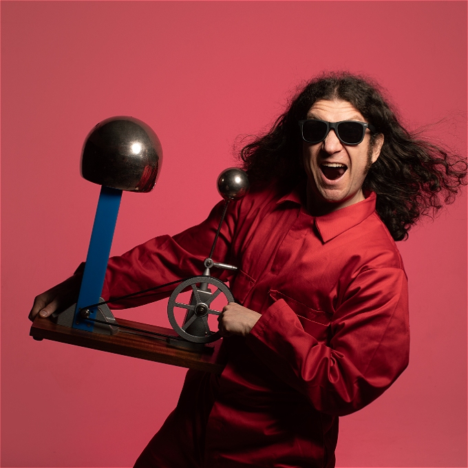 Male comedian with long curly brown hair dressed in a red jumpsuit and sunglasses holding a Van der Graaf Generator