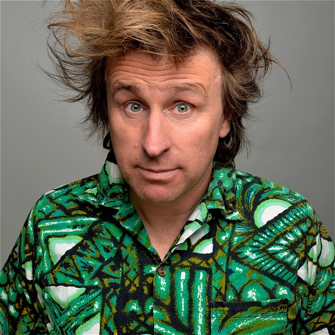 Male comedian with brown hair and a green patterned shirt in front of a grey background