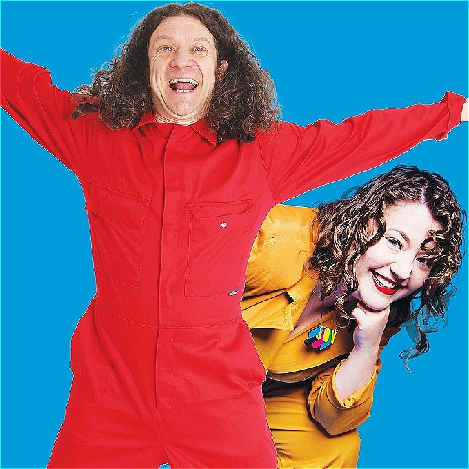 A male comedian in a red jumpsuit with a female comic in a mustard jumpsuit popping her head out from behind him.