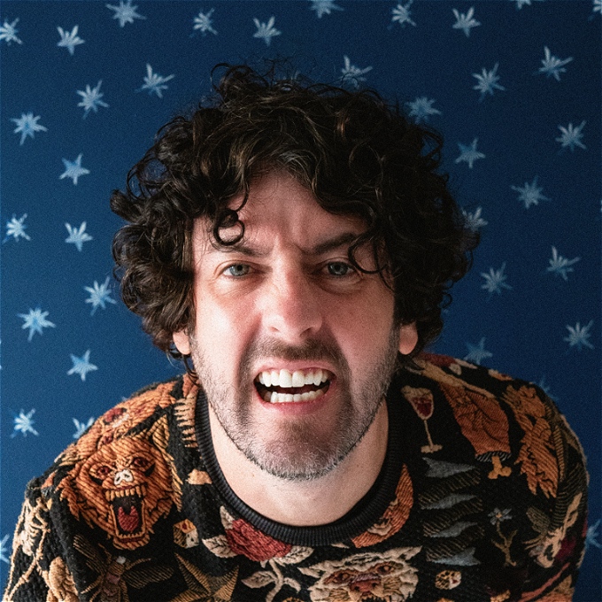 Male comedian, dark curly mid length hair, squinting confused to camera