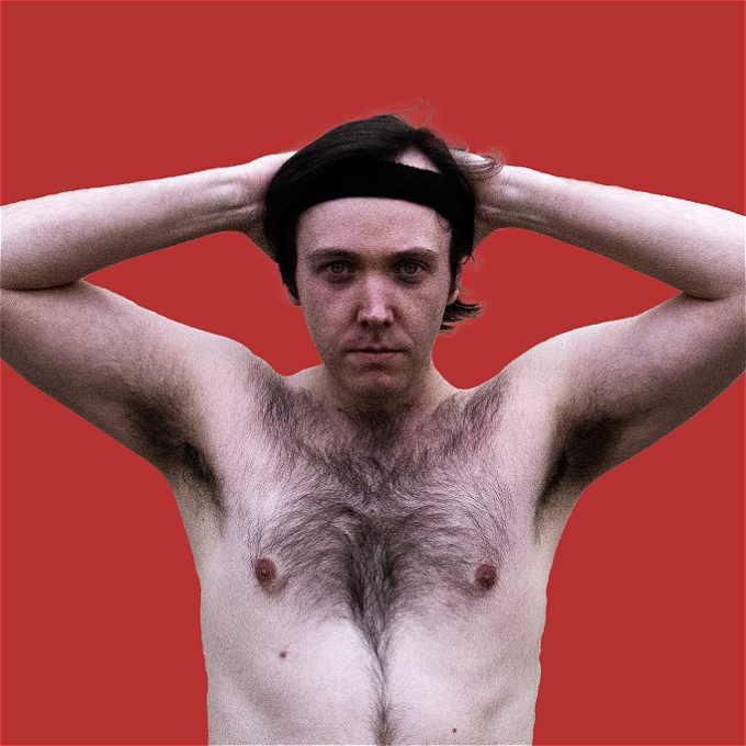 The upper body of a male shirtless comedian with brown hair, swept back and held in place by a sweatband. He has his arms held up behind his head.