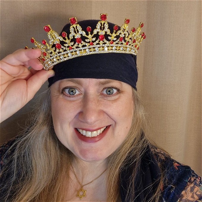 Female comedian with blonde hair, wearing a gold star of David and blue headscarf, is placing a crown on her head