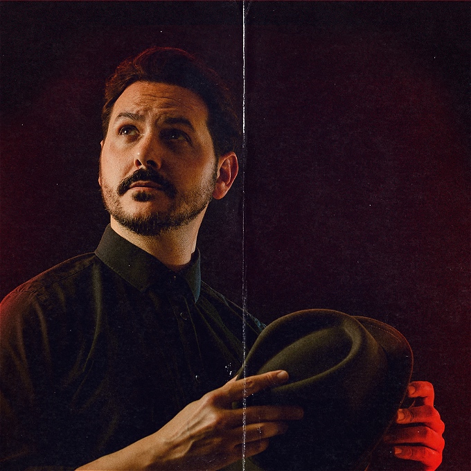 Ignacio Lopez comedian stands in a dark room, looking upwards, holding a fedora in his hands.