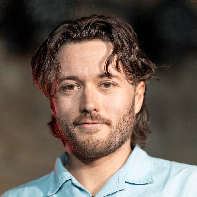 Male beared comedian with longish brown hair and brown eyes wearing a blue shirt