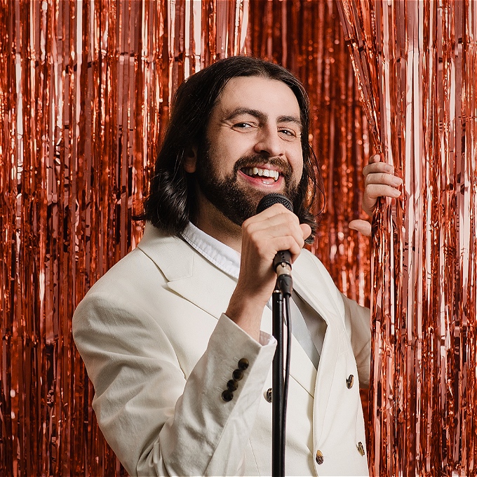 A comedian with long dark hair and a beard, holds a sparkly foil curtain open with one hand and a microphone to his mouth with his other hand. He has a big smile on his face.