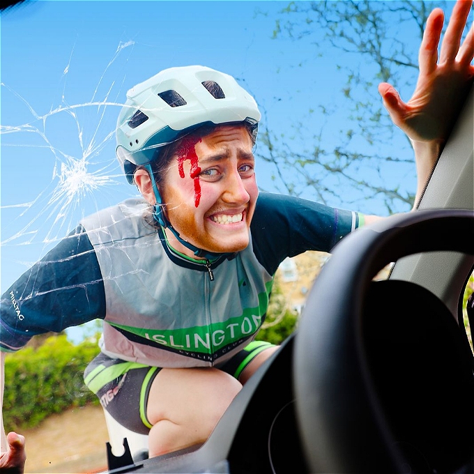 Drag king, character comedian Kathy Maniura dressed in cycling lycra and a helmet on the smashed window of a car with blood on their forehead