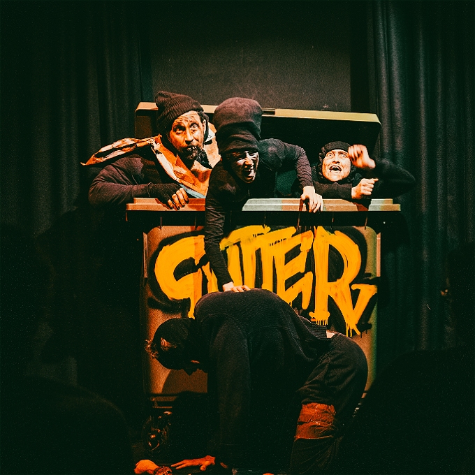 Three bouffon clowns emerge from a big bin with the word 'Gutter' graffitied on the front