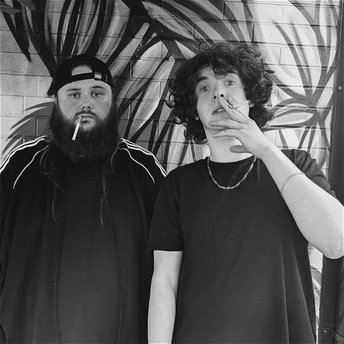 Black and white photo two comedians staring at the camera with cigarettes in their mouths. Ash is in a tracksuit with a big bushy beard and backwards cap. Max is in a black t-shirt with long curly hair. Ash looks quite despondent. Max looks confused.