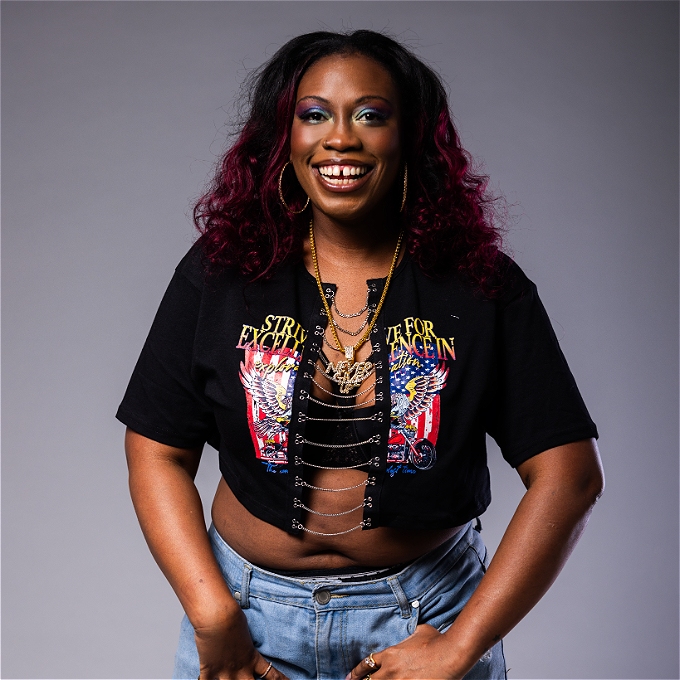 A smiling black woman with long wavy dark hair with red highlights stands against a plain background. She wears a cropped black top with chain detailing, light blue ripped denim shorts, and layered gold jewellery. Her hands rest on her thighs as she poses confidently.