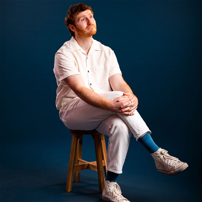 Ali Woods sits casually on a stool in an all white outfit except for blue socks, which match the backdrop he is sat in front of.