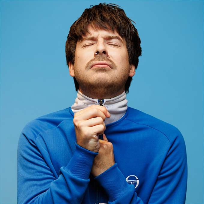 Richard, a white male in his 30s dressed in a cobalt tracksuit jacket stands with his eyes closed in front of a light blue background. He is clutching the zip of his jacket with both hands.