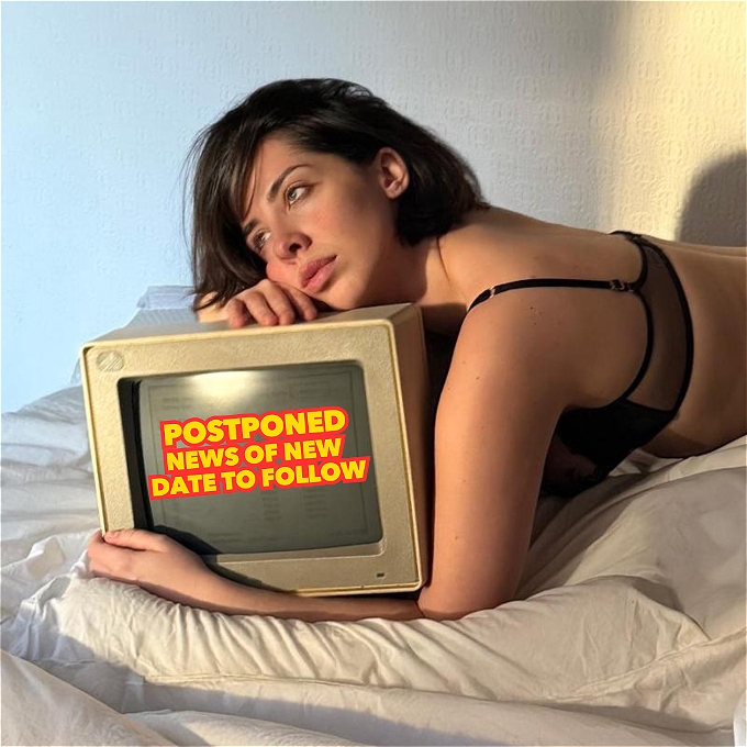 A dark-haired woman looks mournfully out of the window, lying in bed clasping a 90s white desktop computer.