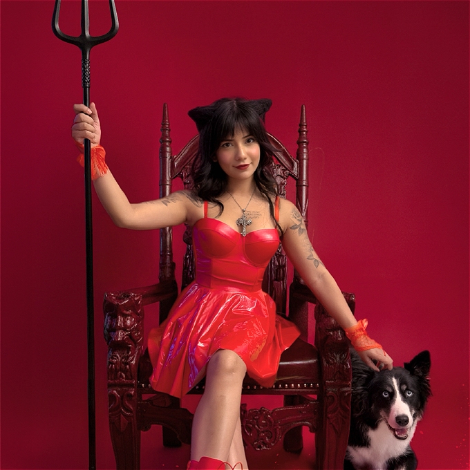 Wearing red PVC Saaniya Abbas sits in a chair holding a trident and stroking a dog