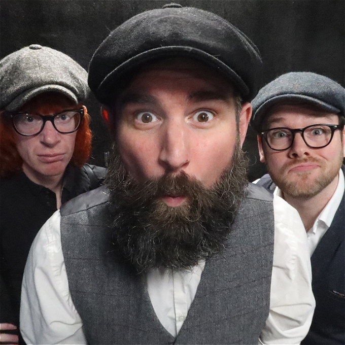 A male musical comedian in flat cap, grey waistcoat and white shirt leans in to stare at the camera with a cheeky look on his face and an impressive beard. Behind him are his band members, also in flat caps, leaning in and staring at the camera, with wry smiles on their faces. They both wear glasses.