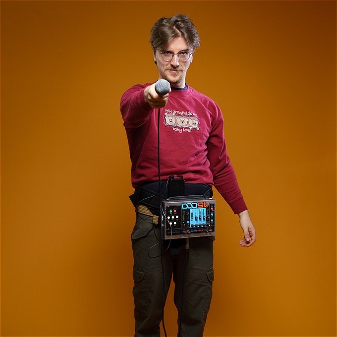 Male comedian with brown hair and glasses with a loop station strapped to his waist, staring straight at you, with a microphone pointing directly at you.