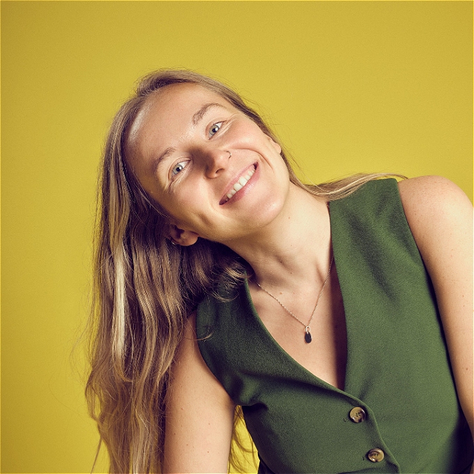 A friendly smiling woman with blonde hair and a green waistcoat. Yellow background. She seems nice.