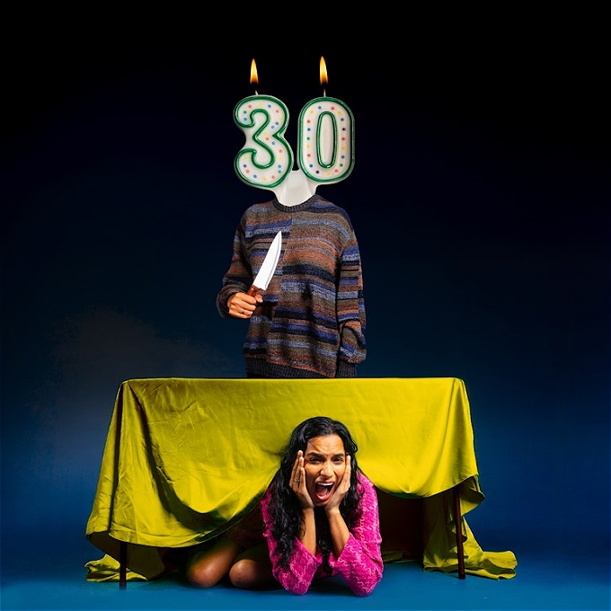 A pretty woman in pink hides under a table with a yellow tablecloth, her hands are clasped on her cheeks and she's screaming. Behind the table is a man's body with a 30th birthday candle for a head, lit, and wielding a kitchen knife.