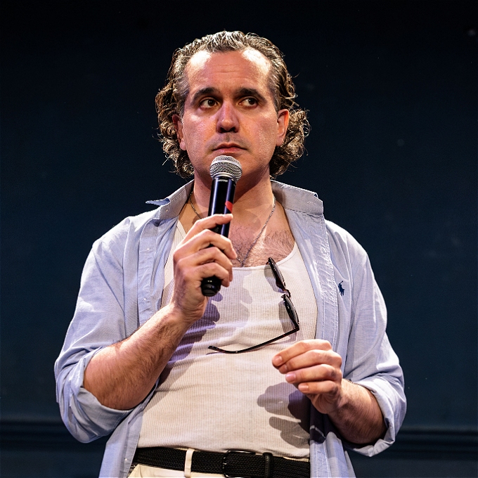 A man with slicked back hair, holding a mic, looking pensively to the side. Wearing a white tank, blue shirt, sunglasses tucked into his shirt.