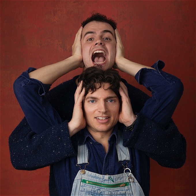 Tall man with a wide open mouth leans his head on another man who half smiles. Their arms are interlocked with hands covering their ears.