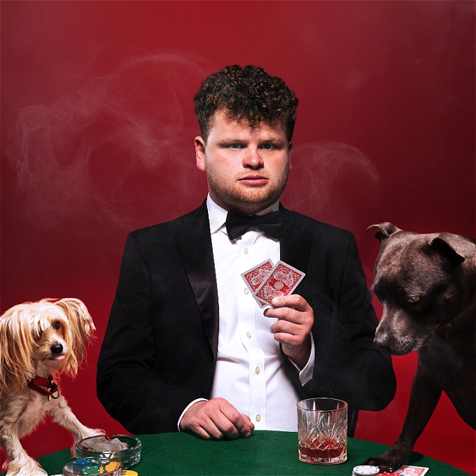 A comedian in a black dinner suit jacket and tiesits at a card table holding two cards. Two dogs also sit at the table. They have their own cards. One also has a whisky, the other. has an ash tray. Smoke bellows across the red background.