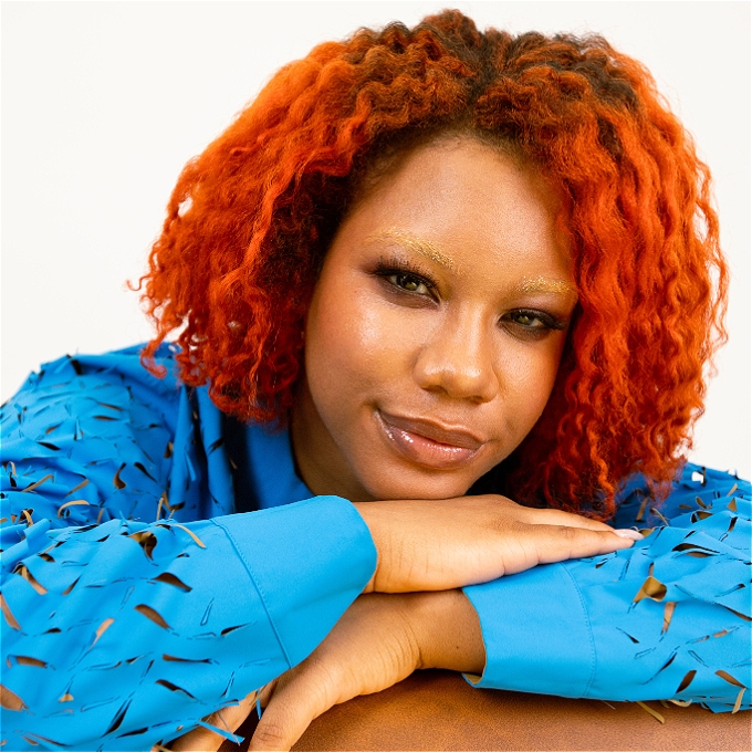 Comedian Kemah Bob, gorgeous caramel skin and tangerine hair, smirks while resting her head on crossed arms.
