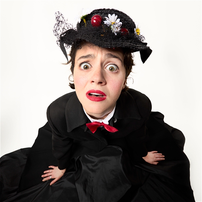 A woman dressed in a white shirt, red bowtie, black coat and black hat with cherries and flowers. She looks frustrated, or confused.