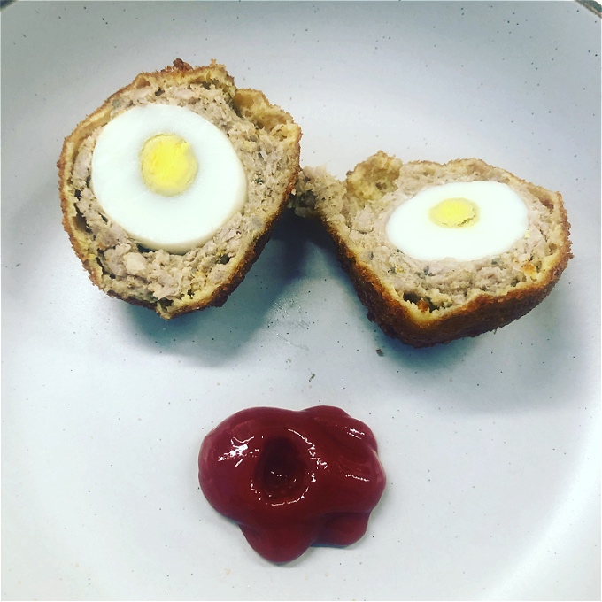 Photograph of a plate with the Scotch egg chopped in half and a dollop of tomato sauce. They have been arranged to make it look like a face.