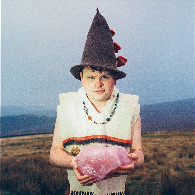 A male comedian wears a pointy hat and holds a large piece of rose quartz while standing on a moorland on a grey day.