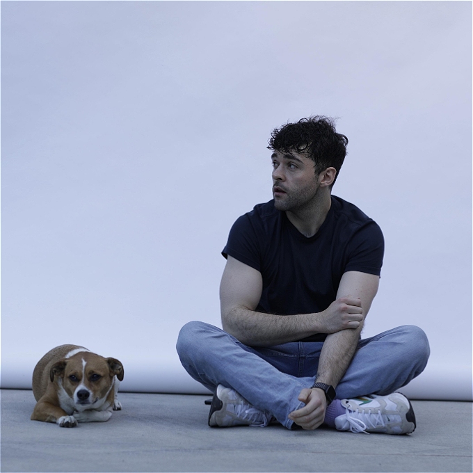 Male comedian with black hair sitting on floor next to dog