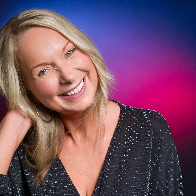 Female comedian with blonde hair and a black sparkly top in front of a pink and blue background