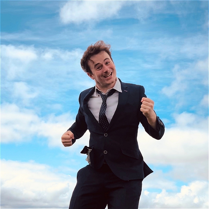 Alex Kitson (young, dark-haired male) is smiling gleefully at the camera in the foreground. In the background is a bright blue sky and village.