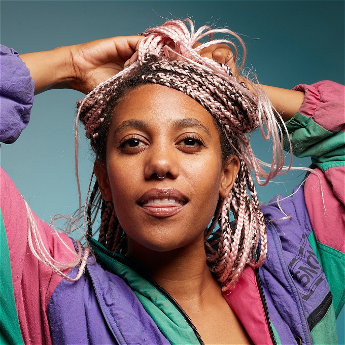 Comedian Kate Cheka, wearing a snowsuit, with her arms up and in her pink hair.